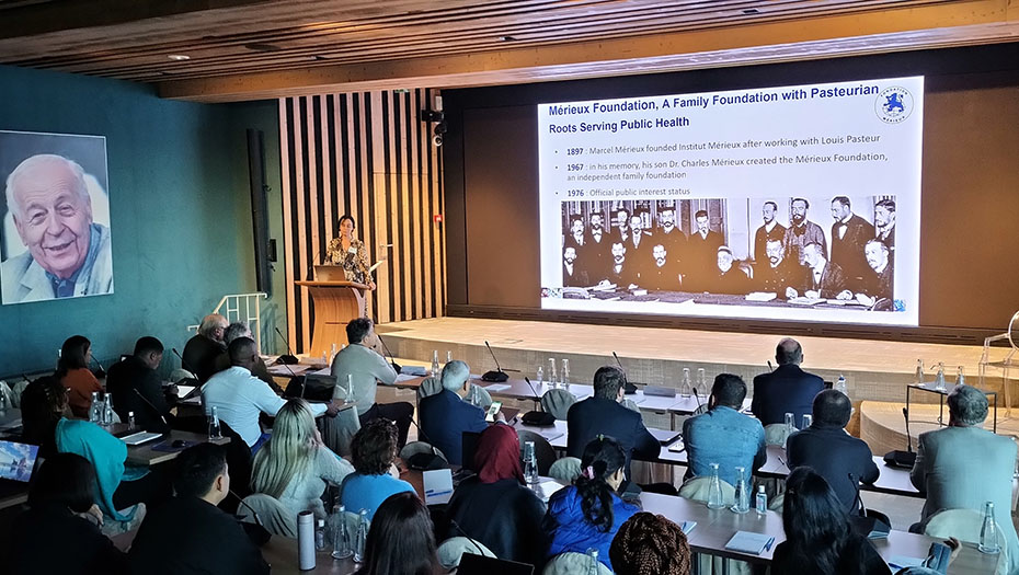 Une présentation sur la Fondation Mérieux avec une personne sur le podium devant un public. Un grand portrait est accroché au mur de gauche.