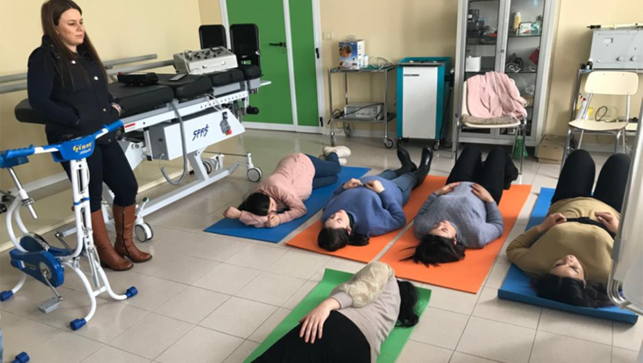 A group of women lying on floor mats in a room, guided by a standing instructor.