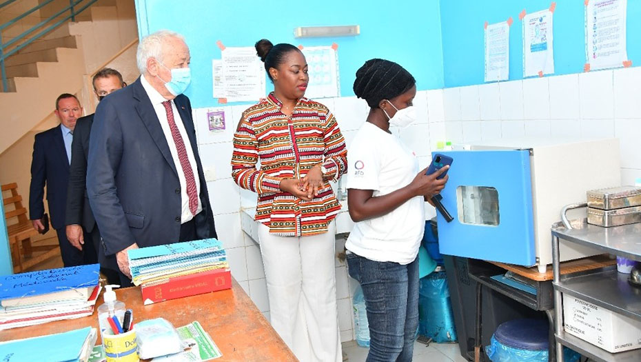 An official delegation visits a laboratory, with a female technician demonstrating medical equipment.