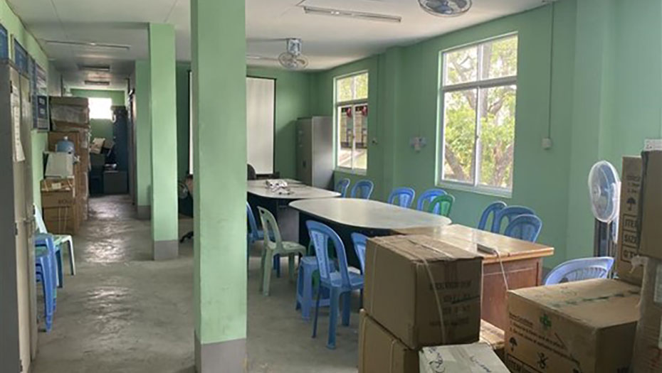 A laboratory room before renovation, with green walls, blue plastic tables and chairs, and stacked cardboard boxes.