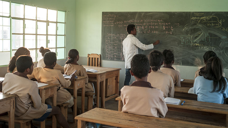 A teacher writes on a blackboard in front of students seated at wooden desks in a classroom.