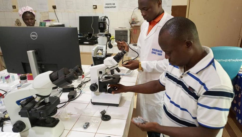 Two laboratory technicians work on microscopes in a laboratory, with computers and various scientific equipment on the tables.