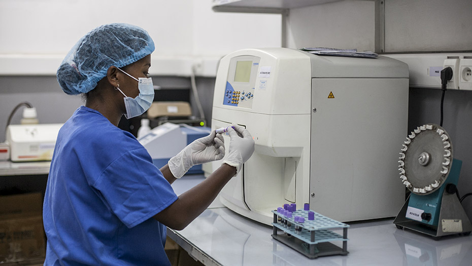 Une technicienne de laboratoire en blouse bleue, masque et charlotte utilise une machine de diagnostic avec des échantillons de sang.