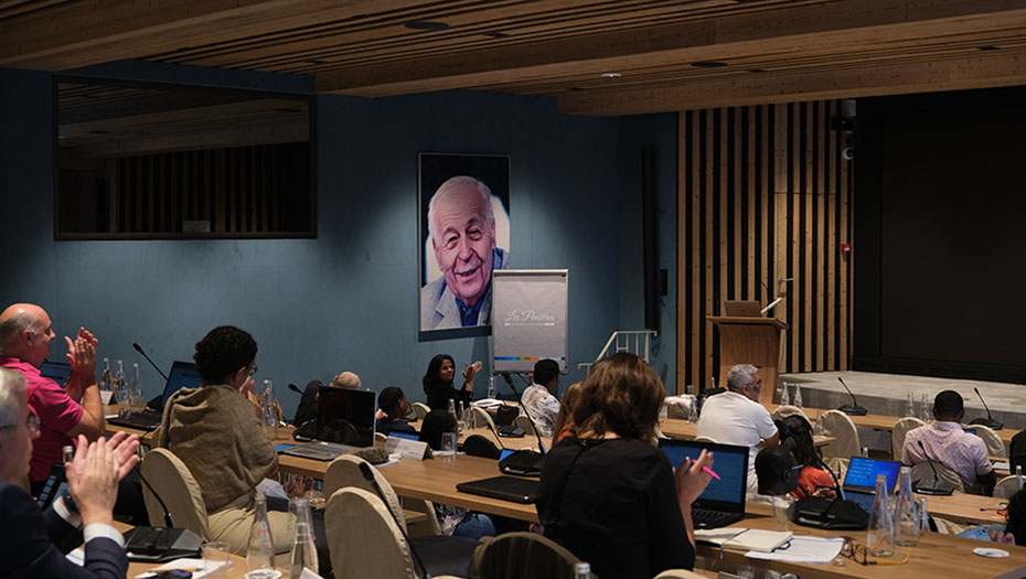 Participants applaud during a conference in a room at the Center des Pensières, with a large portrait on the wall and laptops on the tables.