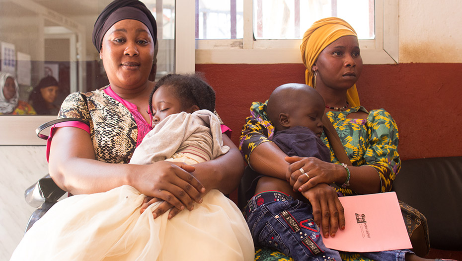 Two women sitting side by side in a waiting room, each holding a sleeping child in their arms, with other people in the background.