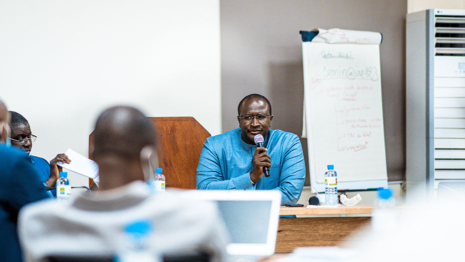 A man sitting at a table, speaking into a microphone during a class, with a whiteboard in the background and participants.