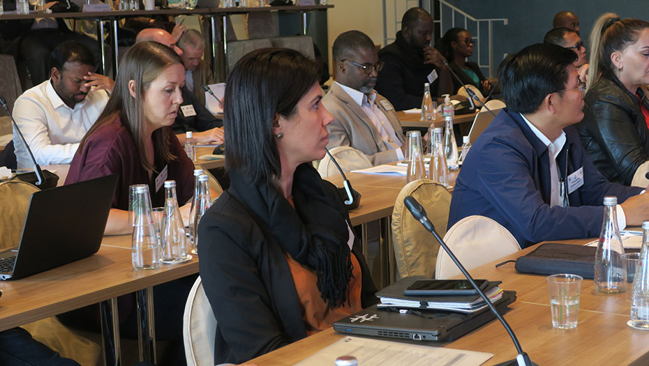 Participants assis à des tables avec ordinateurs et carnets dans une salle de conférence équipée de micros et d’un pupitre.