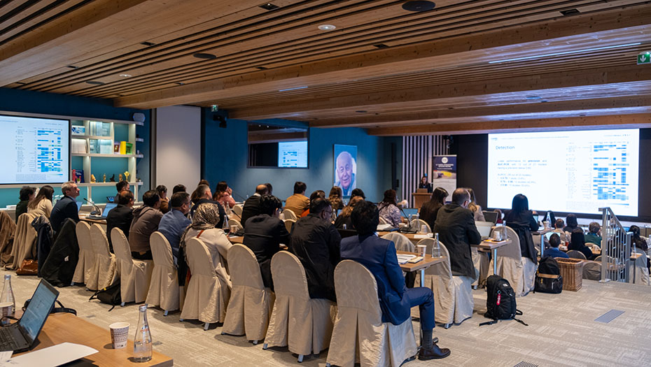 Participants assis dans une salle de conférence avec deux écrans affichant des présentations, certains utilisant des ordinateurs portables.