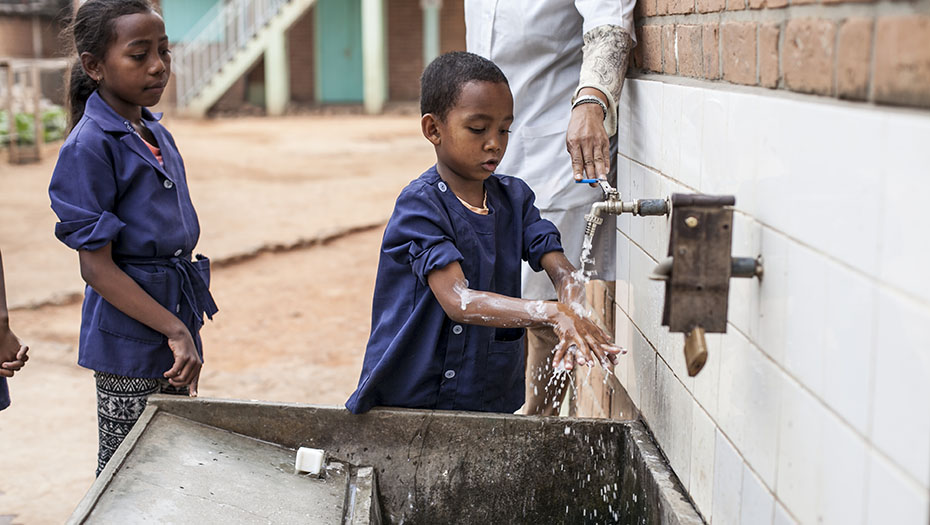 Child washing hands under an outdoor tap, assisted by an adult and other children, in front of a tiled wall with a basin