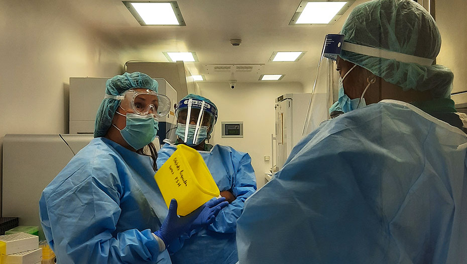 Trois personnes en tenue de protection complète dans un laboratoire, dont une tenant un conteneur jaune de déchets médicaux.