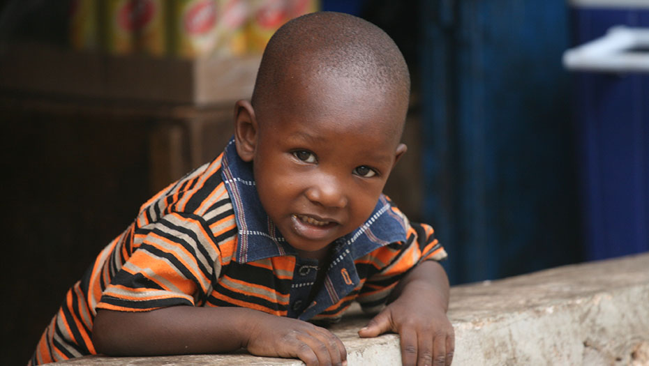 Jeune enfant en tee-shirt rayé appuyé sur un muret, avec des boîtes empilées et une porte bleue en arrière-plan.