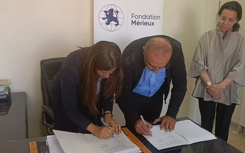 Two people signing documents on a table in front of a logo panel, a third person standing nearby observing.