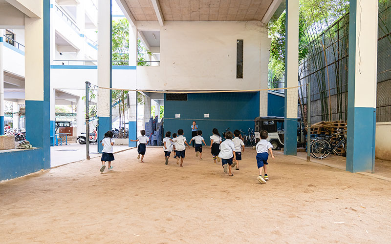 Children running under an open building with a dirt floor and a net suspended in the center.