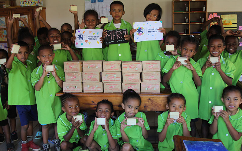 Group of children wearing green uniforms holding soap bars in front of stacked boxes on a table in a classroom.