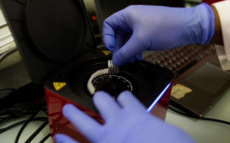 Gloved hand placing tubes into an open laboratory device with screen and keyboard in the background.