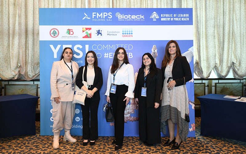 Group of people posing in front of a banner with logos during an indoor event with tables and posters.