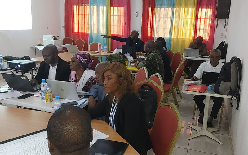 Participants seated with laptops and documents in a training room with tables and colorful curtains.