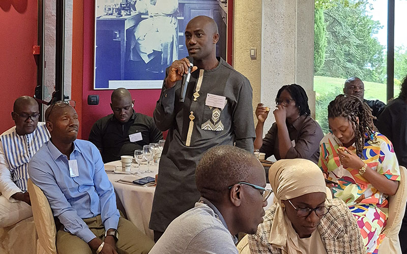 Participant standing with a microphone speaking to a group seated around tables during a workshop.