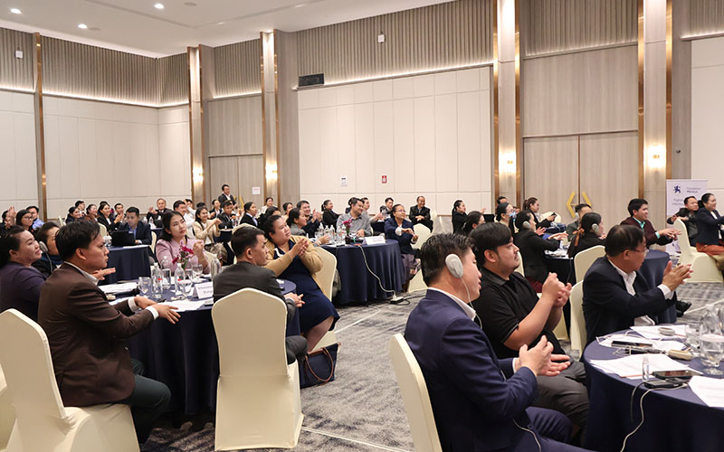 Participants seated in a conference room around round tables, applauding and using audio equipment during an event.