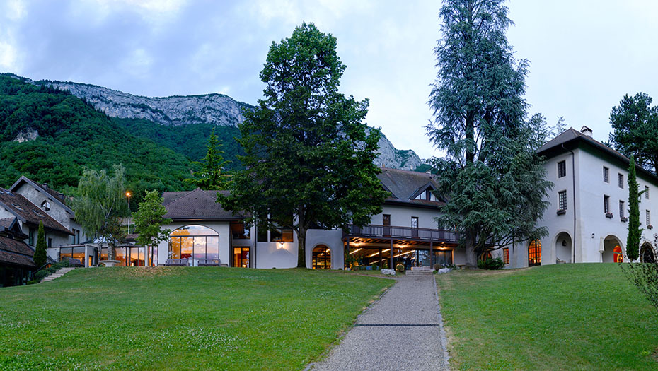 Building with large windows and a lawn in the foreground at the foot of a wooded mountain.