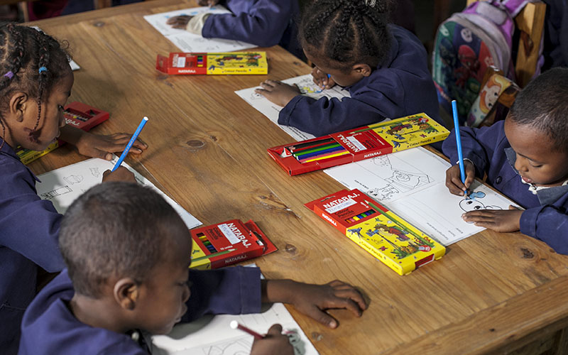 Children seated around a wooden table drawing with colored pencils on paper sheets, surrounded by boxes of school supplies.