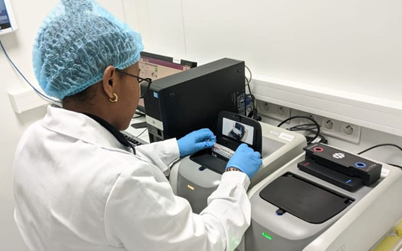 Person in lab coat, gloves and cap inserting a sample into a laboratory analyzer.