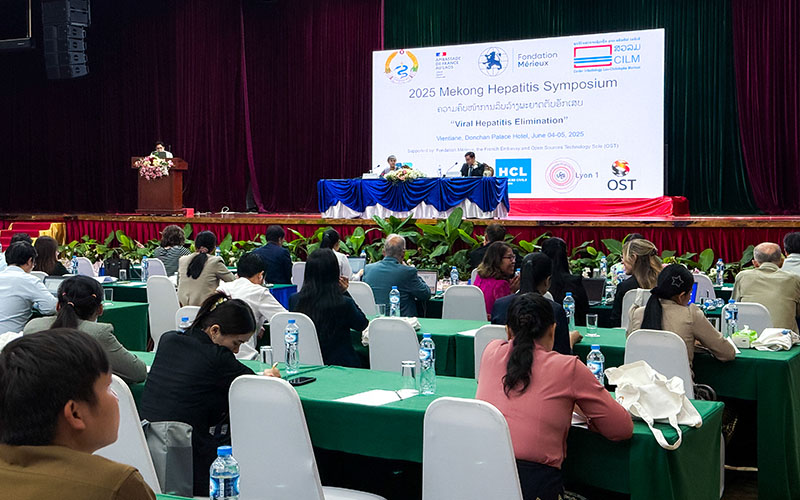 Participants seated at tables with laptops and water bottles facing a stage with a screen during a conference.